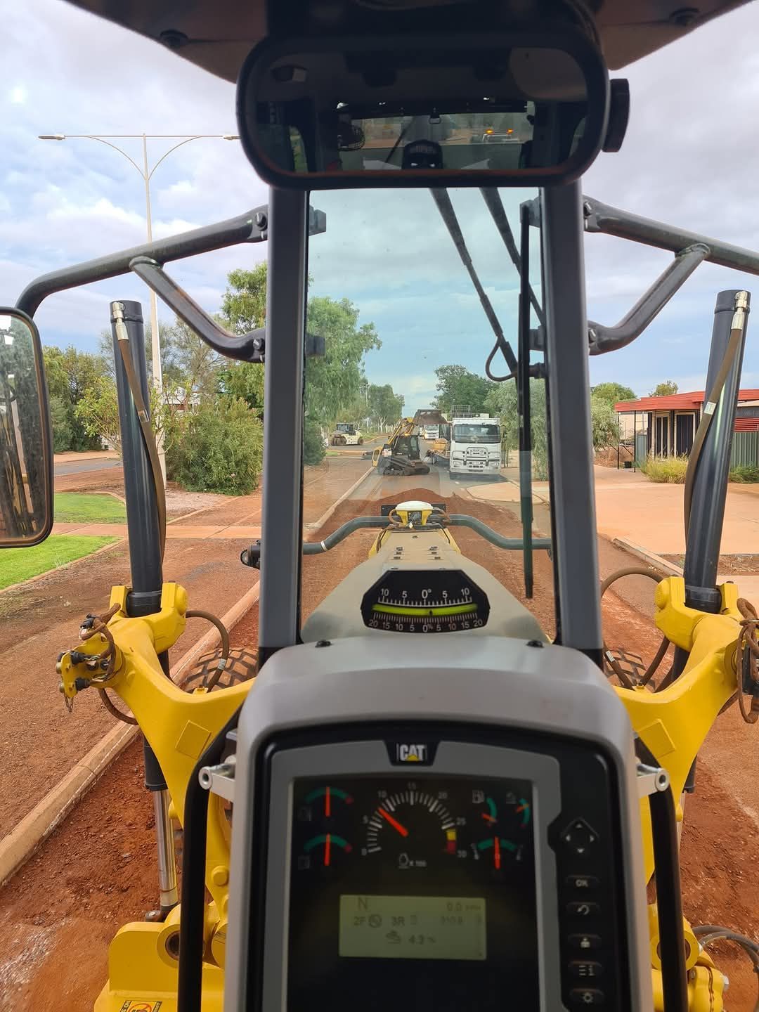 View from inside a yellow grader, looking down a dirt road with other vehicles.