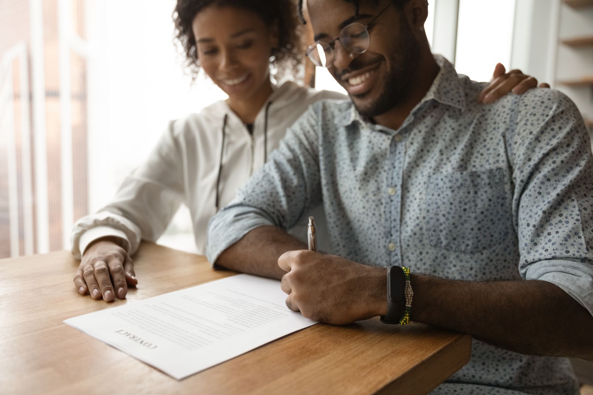 Man signing a document at a table, smiling while woman puts hand on his shoulder.