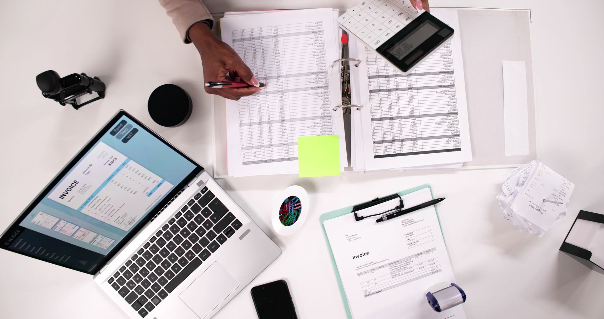 Overhead view of a person's hands working on paperwork at a desk with a laptop, calculator, and other office supplies.