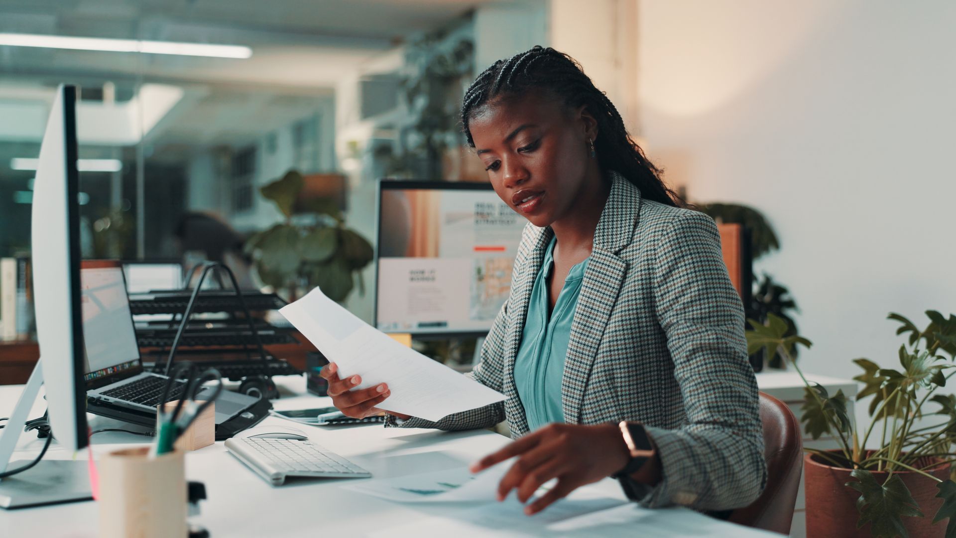 Woman reviews documents at her office desk with a computer and potted plant.