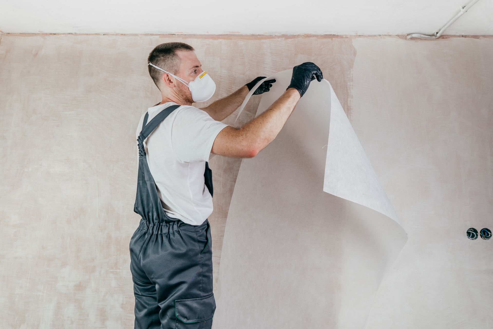 A man wearing a mask and gloves is peeling wallpaper from a wall.