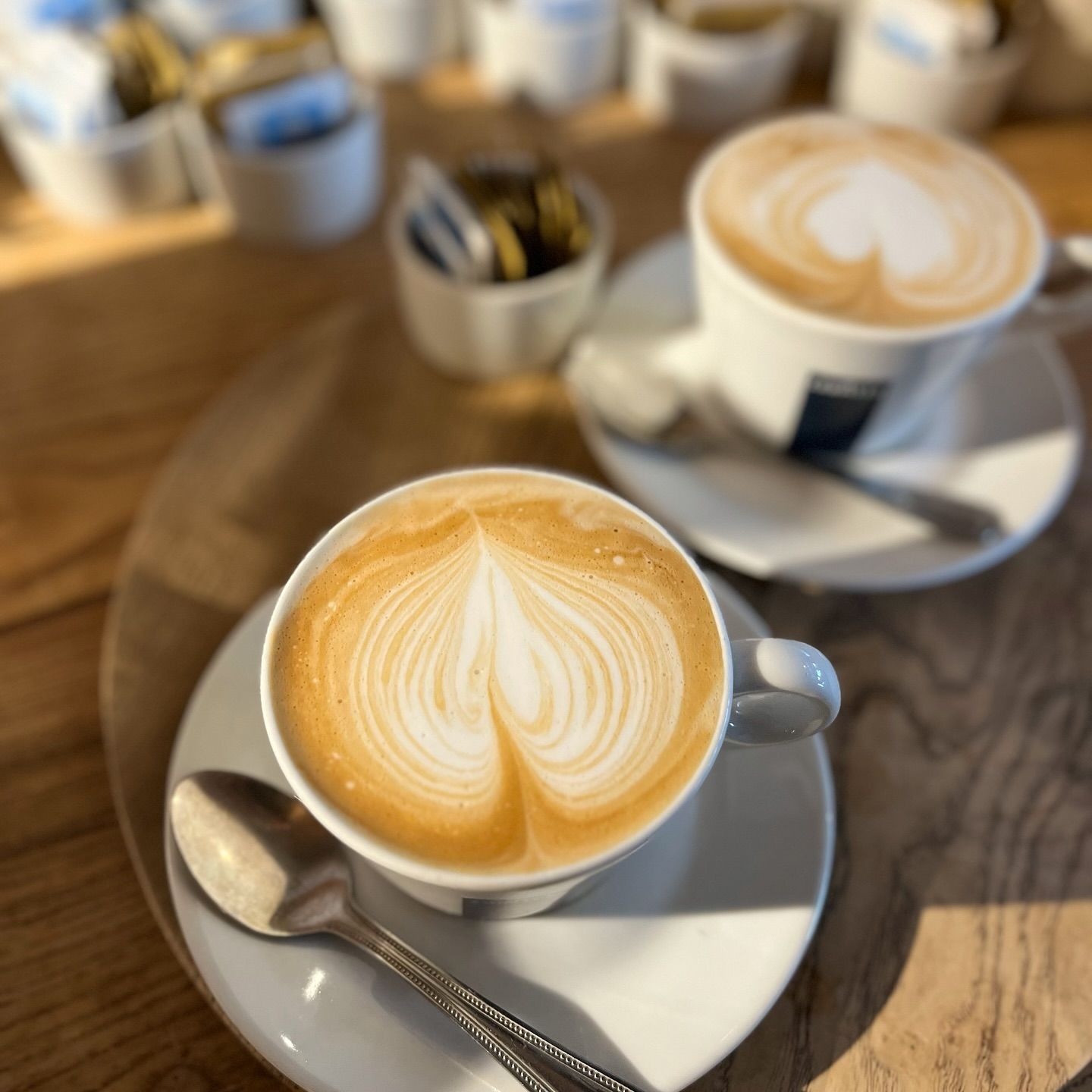 Two lattes on saucers with heart-shaped foam art; spoons rest nearby, set on a wooden table.