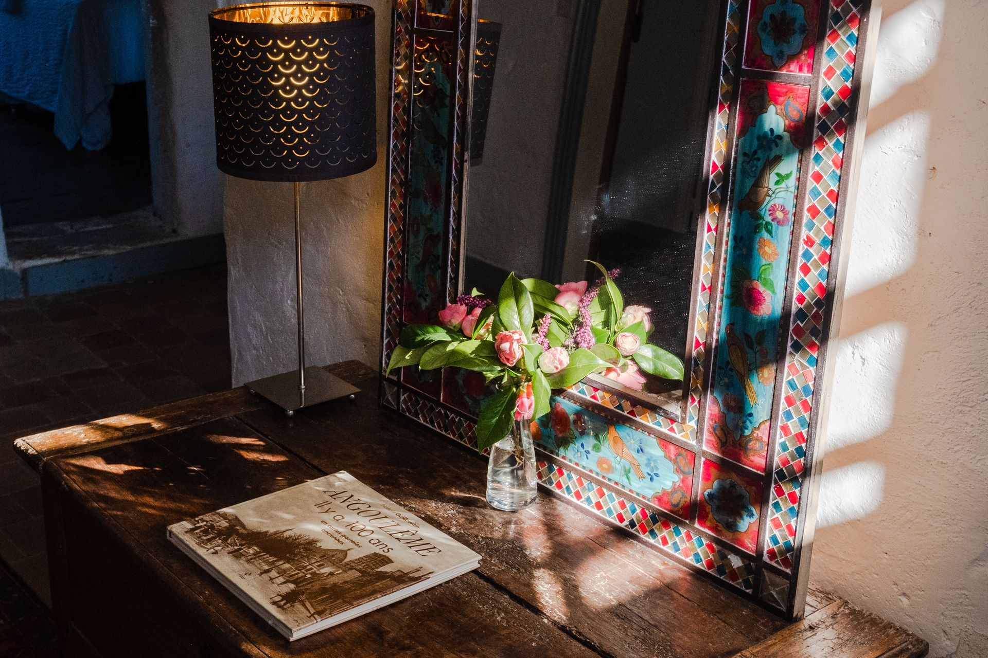 Une table avec un miroir, une lampe, un vase de fleurs et un livre dessus.