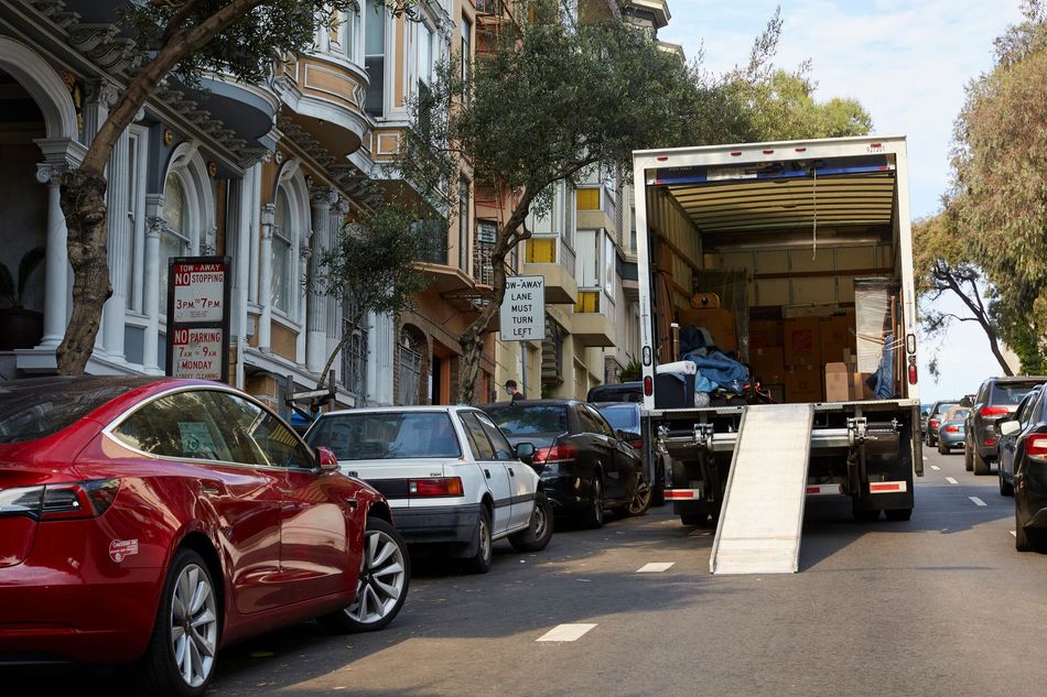 Moving truck with ramp parked on a tree-lined street beside parked cars and buildings
