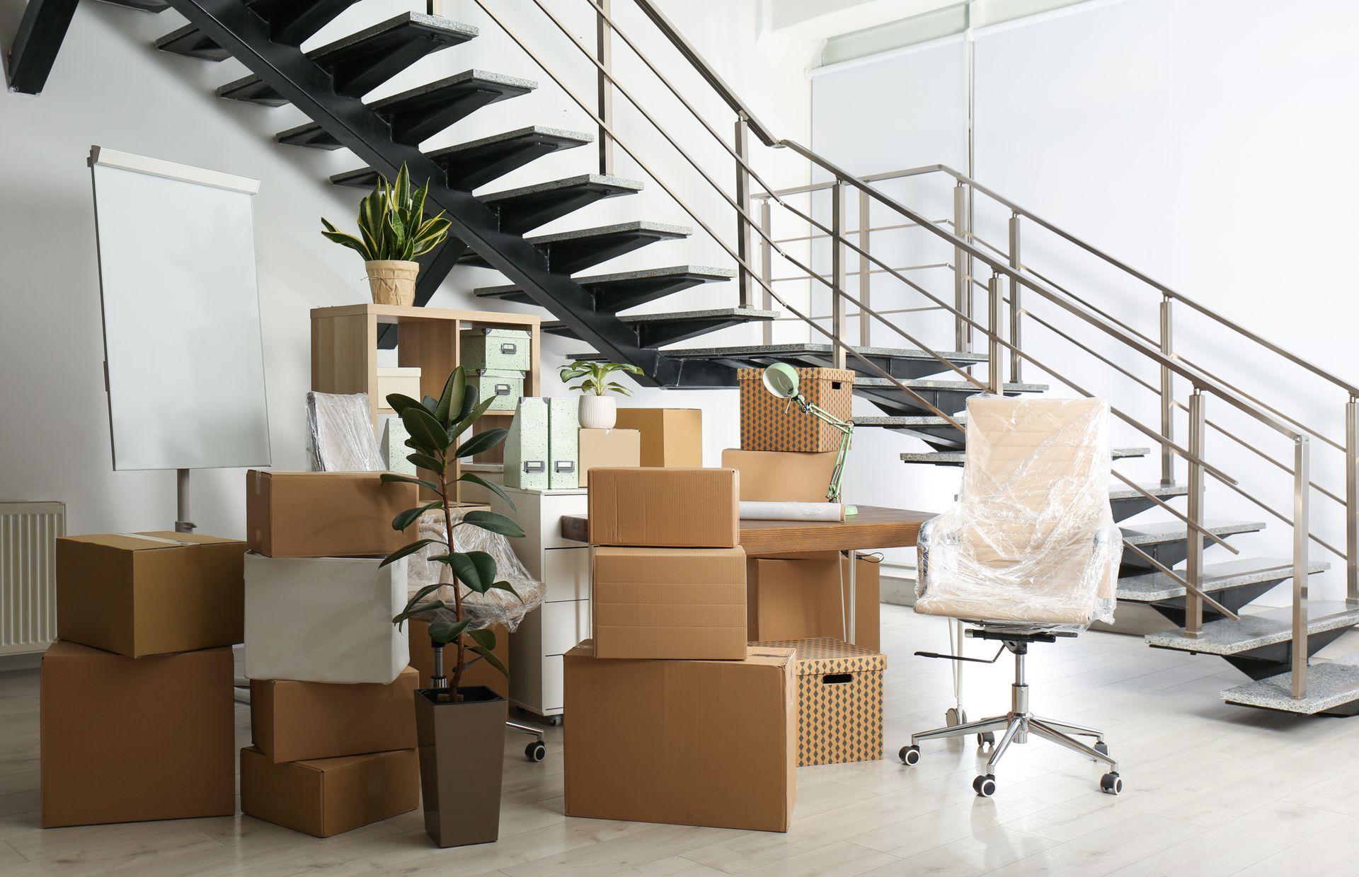 Stacked moving boxes in a bright office with a staircase, chair, and potted plant Stacked moving boxes in a bright office with a staircase, chair, and potted plant