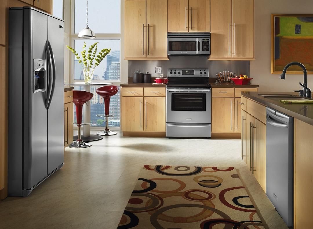 A modern kitchen with light wood cabinets, stainless steel appliances, a patterned rug, and a small dining nook by a window.