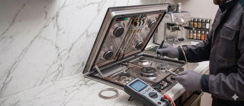 A technician in work clothes uses a multimeter to repair an open stovetop in a kitchen with a marble backsplash.