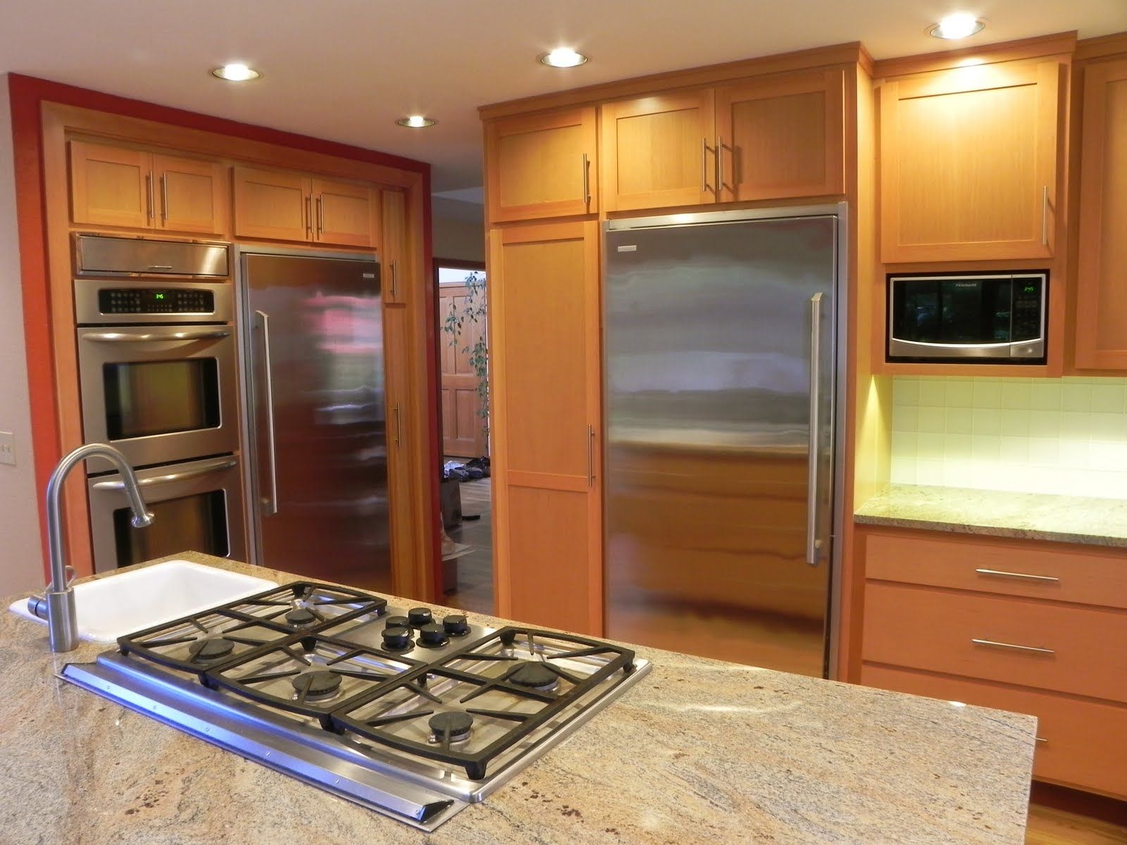 A modern kitchen featuring a granite-topped island with a gas cooktop, stainless steel appliances, and wood cabinetry.