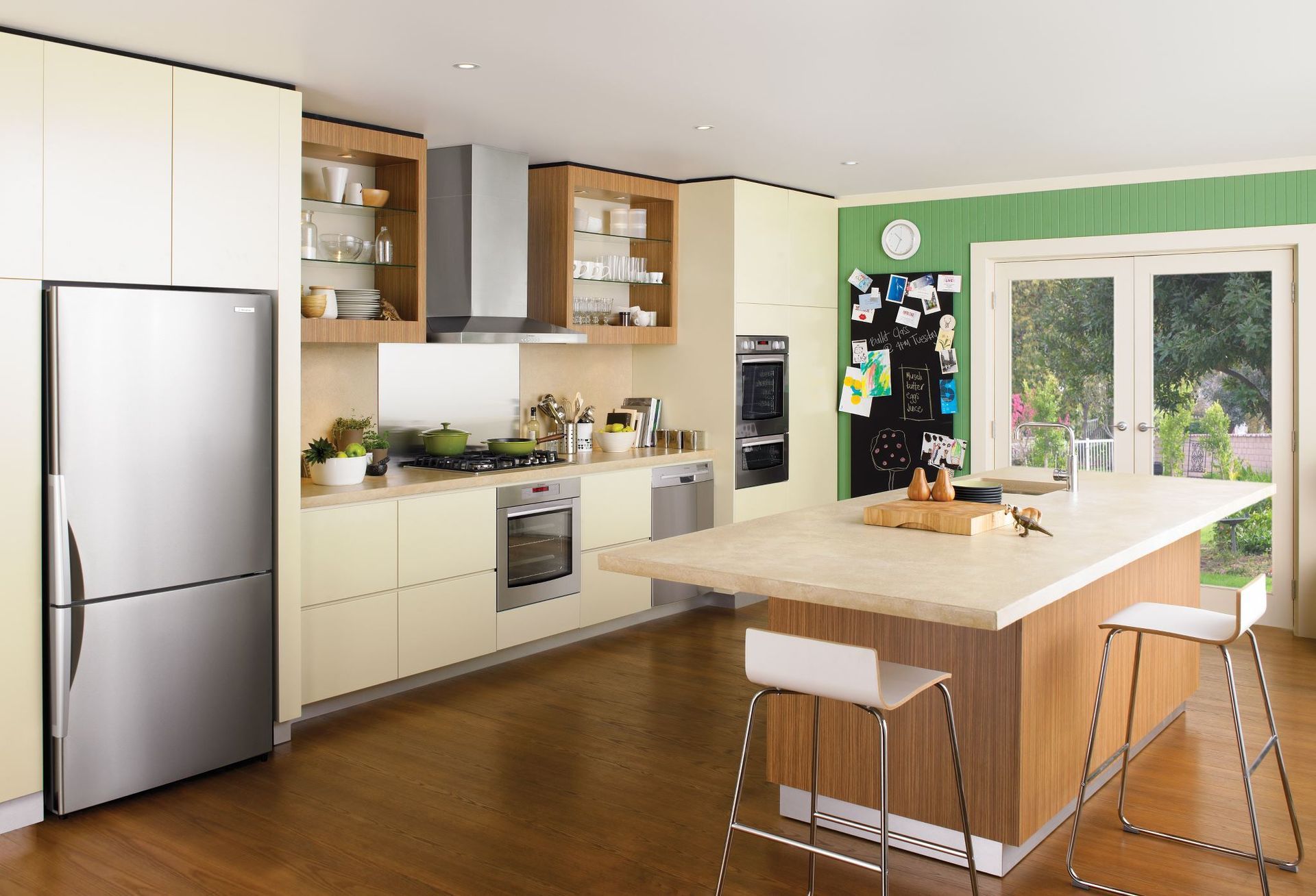A modern kitchen with light yellow cabinets, stainless steel appliances, a wood-topped island, and a green accent wall.
