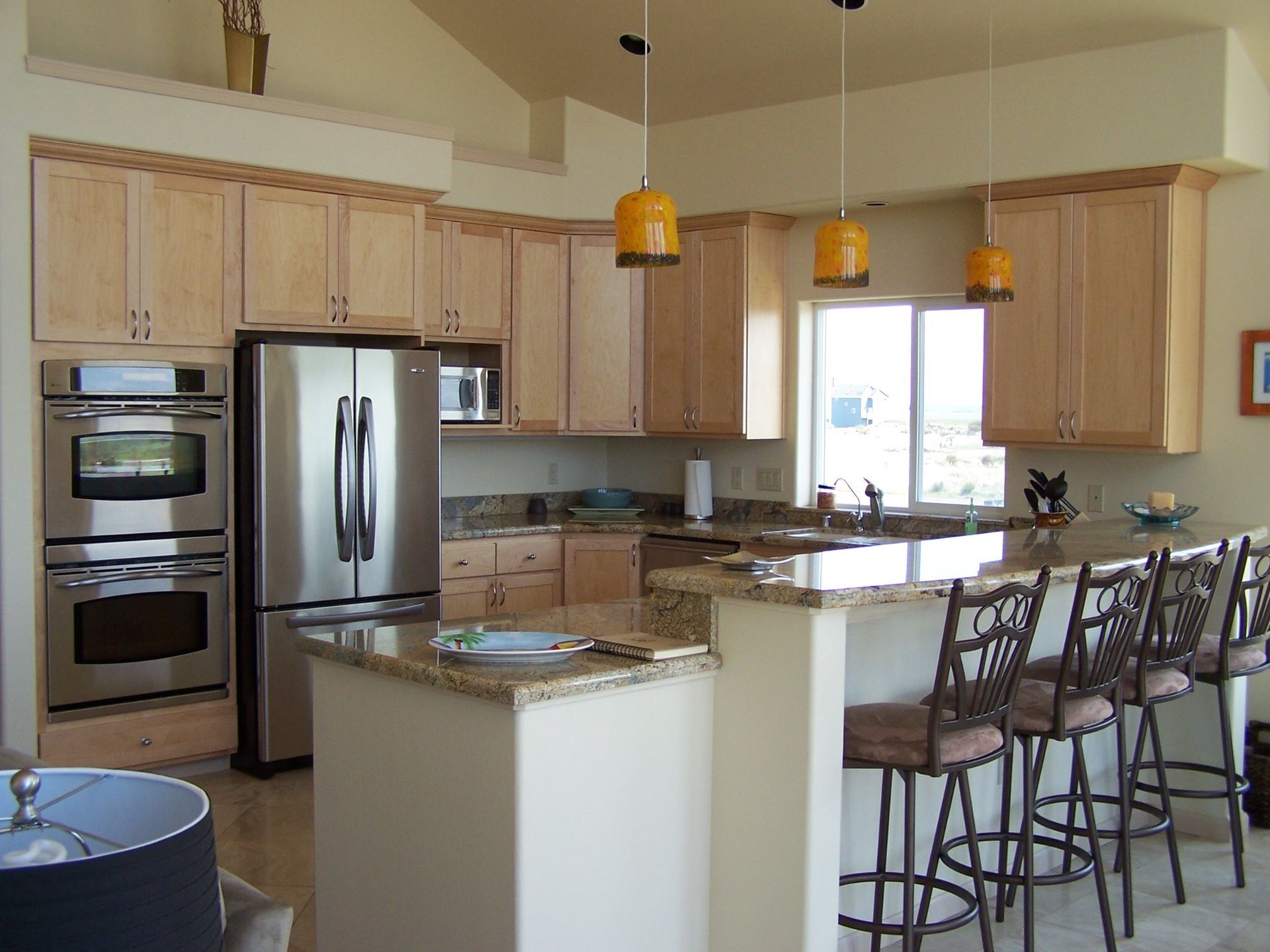 A modern kitchen with light wood cabinets, stainless steel appliances, granite countertops, and a breakfast bar with stools.
