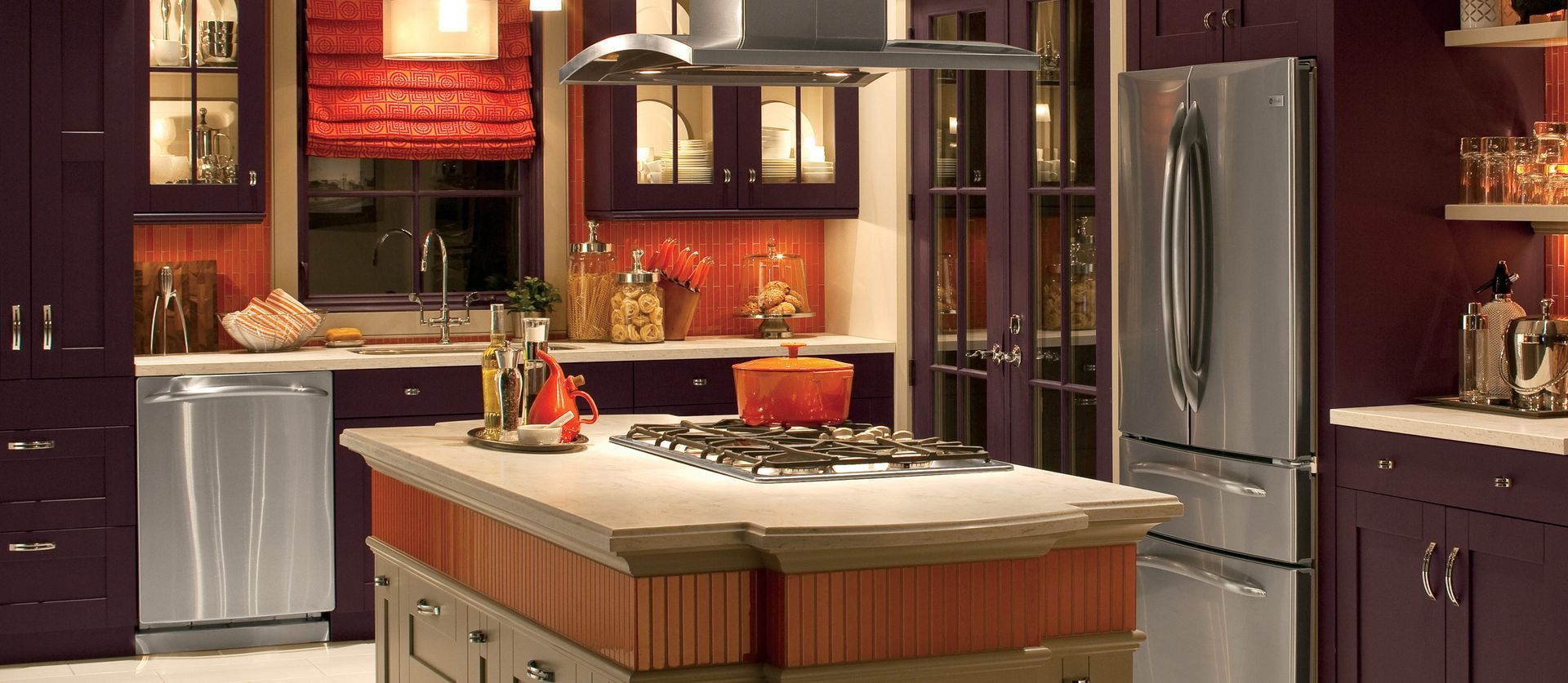 A modern kitchen featuring deep purple cabinetry, stainless steel appliances, a kitchen island, and a reddish window shade.