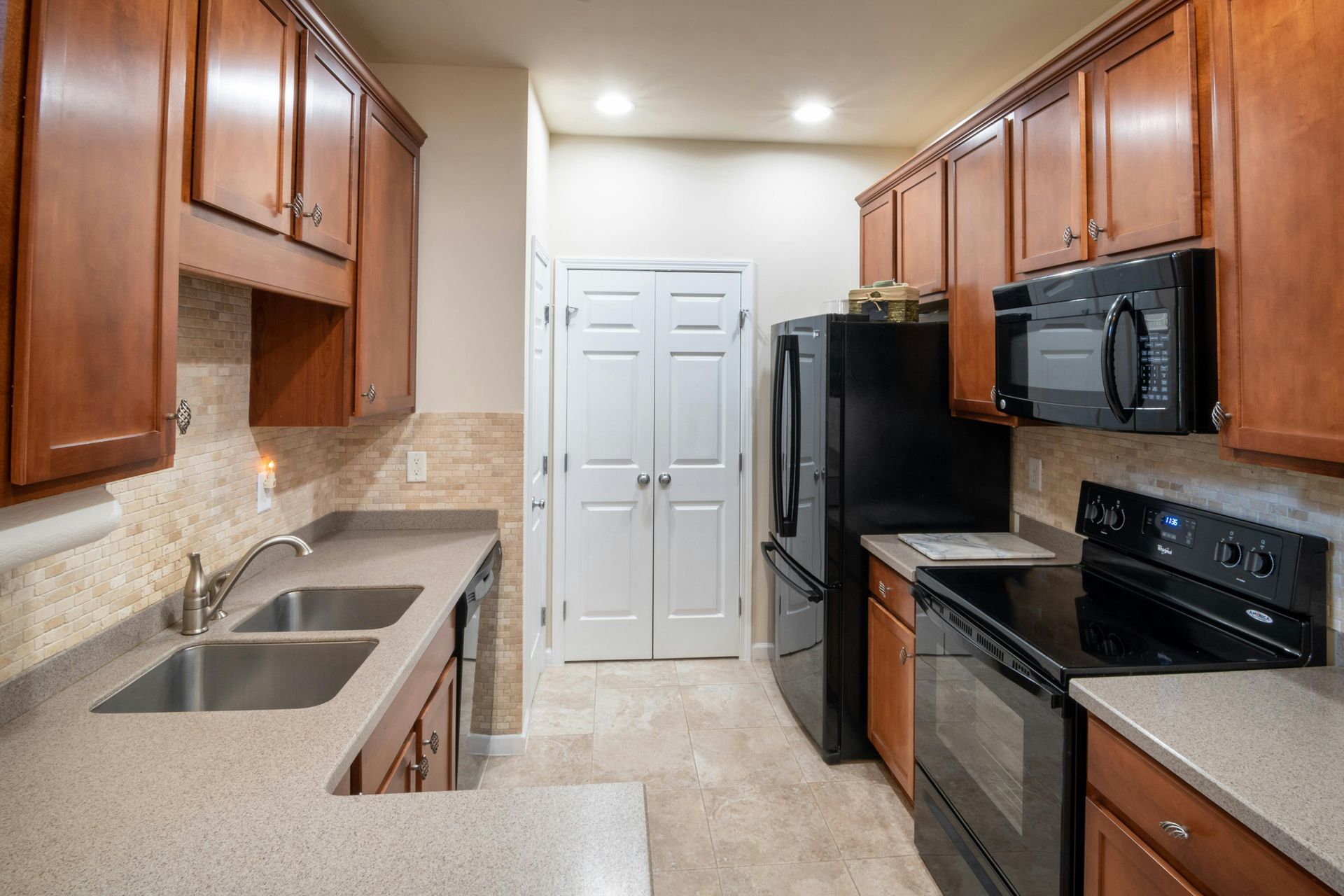 A kitchen featuring light-toned stone countertops, wooden cabinets, stainless steel sink, and black appliances.