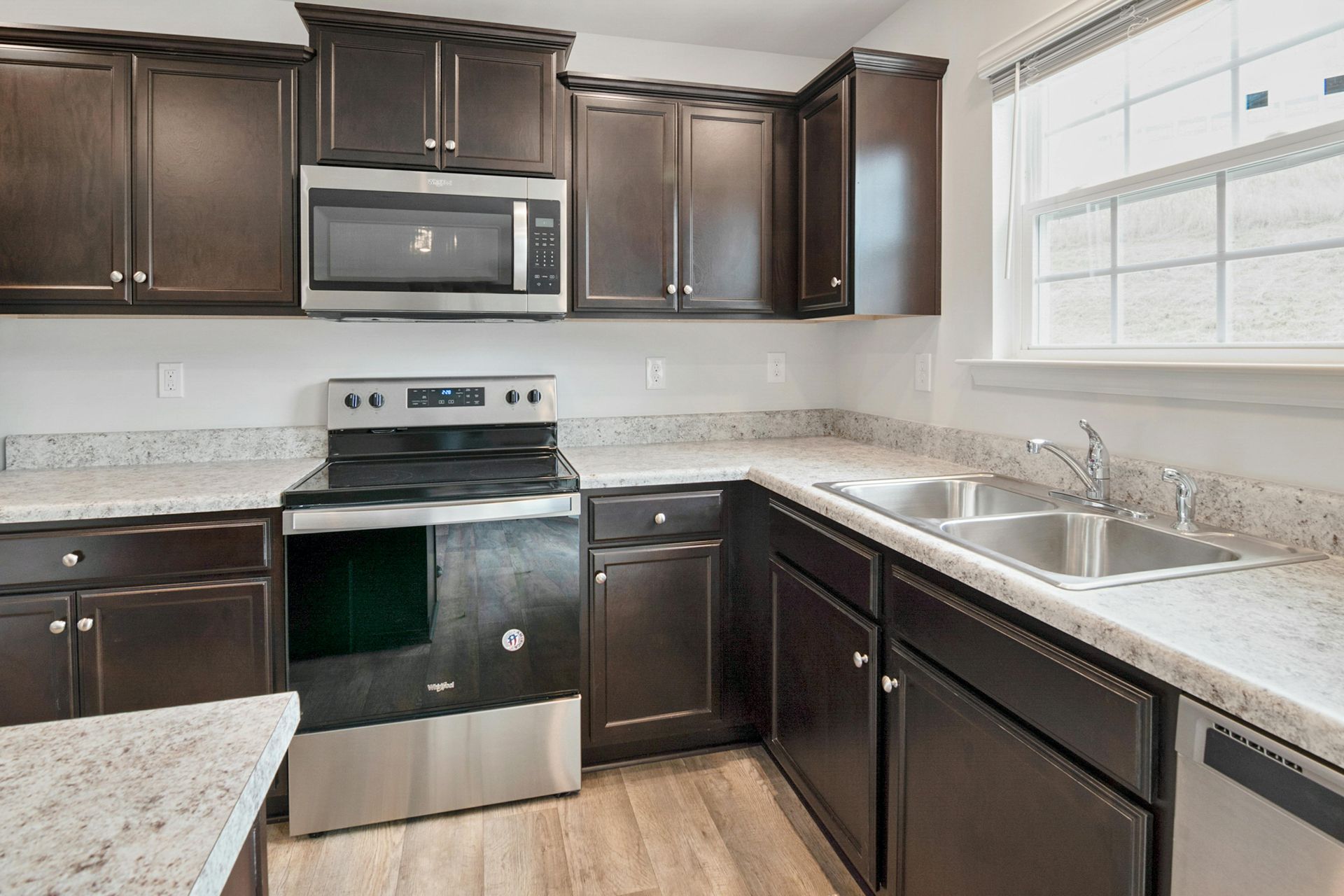 Modern kitchen featuring dark wood cabinets, light granite countertops, stainless steel appliances, and wood-style flooring.