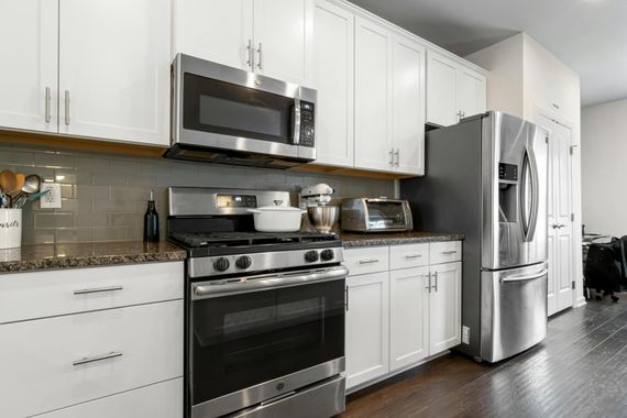 A modern kitchen with white cabinets, stainless steel appliances, a gray tiled backsplash, and dark wood flooring.
