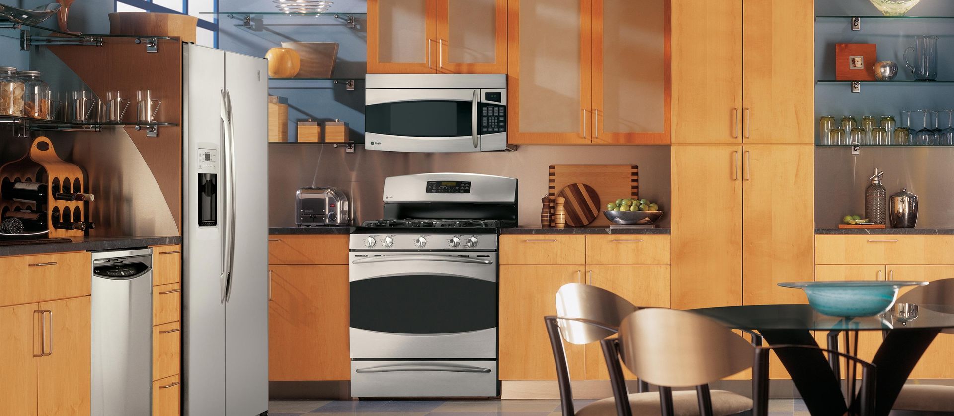 A modern kitchen featuring light wood cabinets, stainless steel appliances, and a small dining table with chairs.
