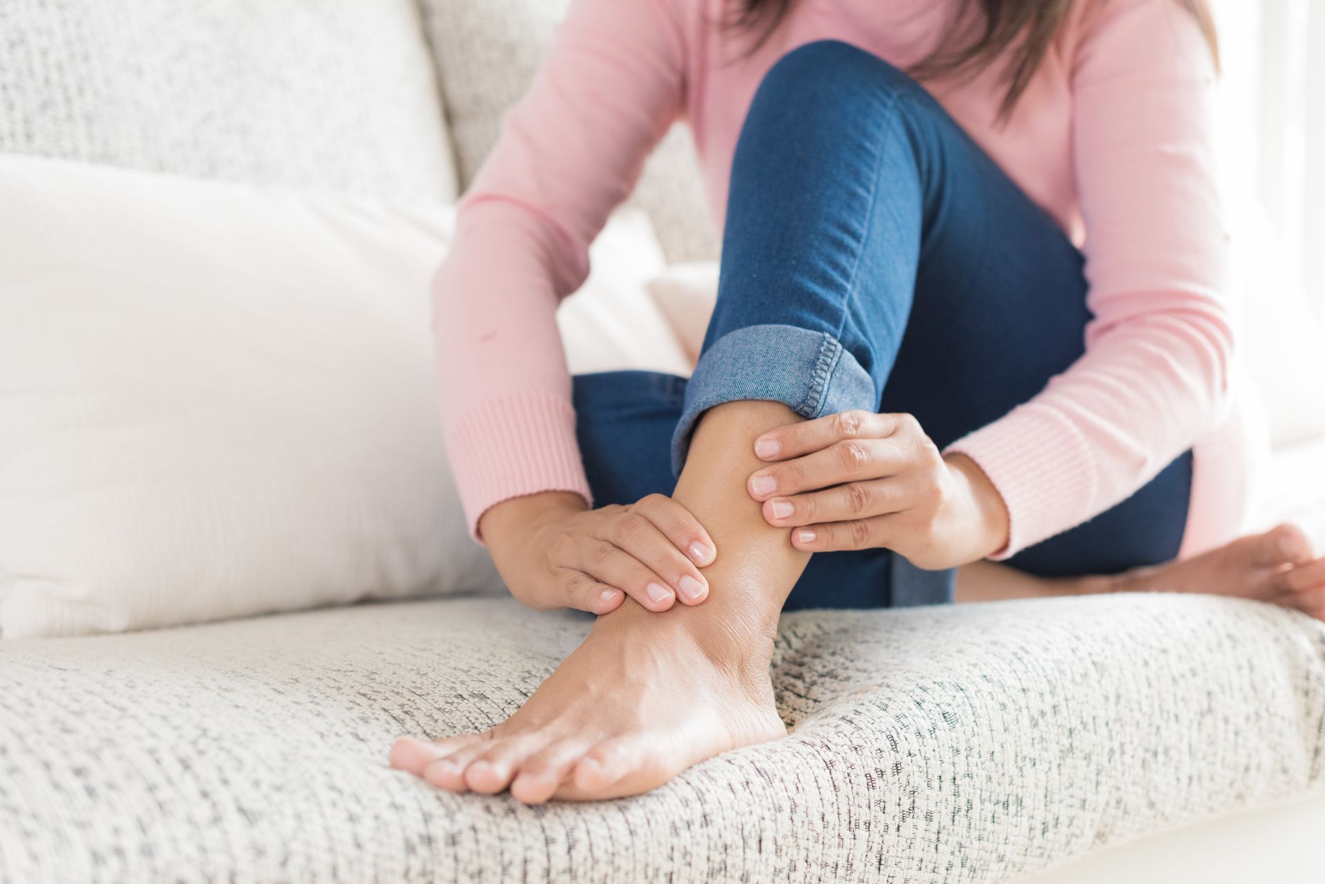Woman seated on a couch, holding her ankle, possibly in pain.