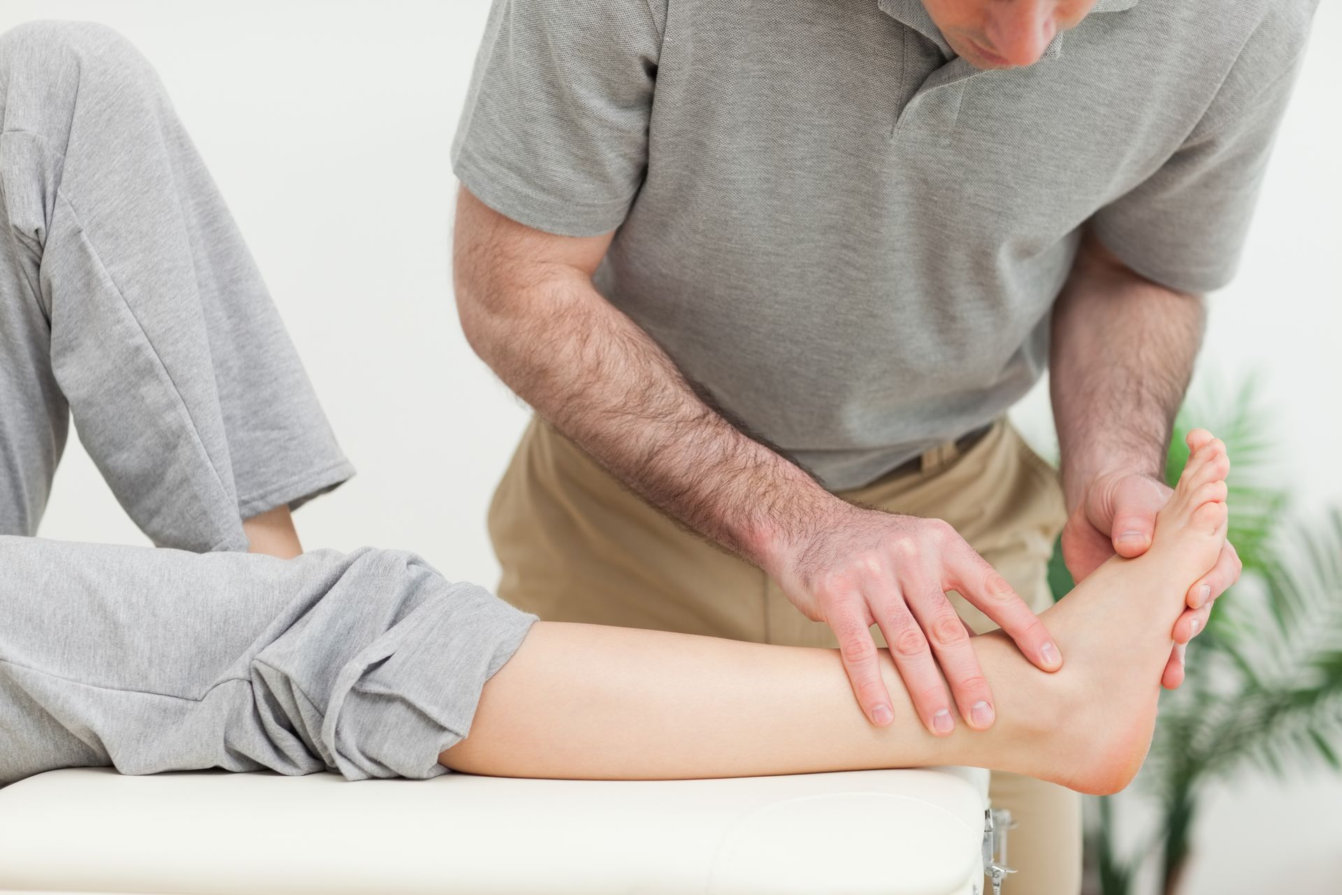 Person examining another person's ankle on a medical table; indoors.