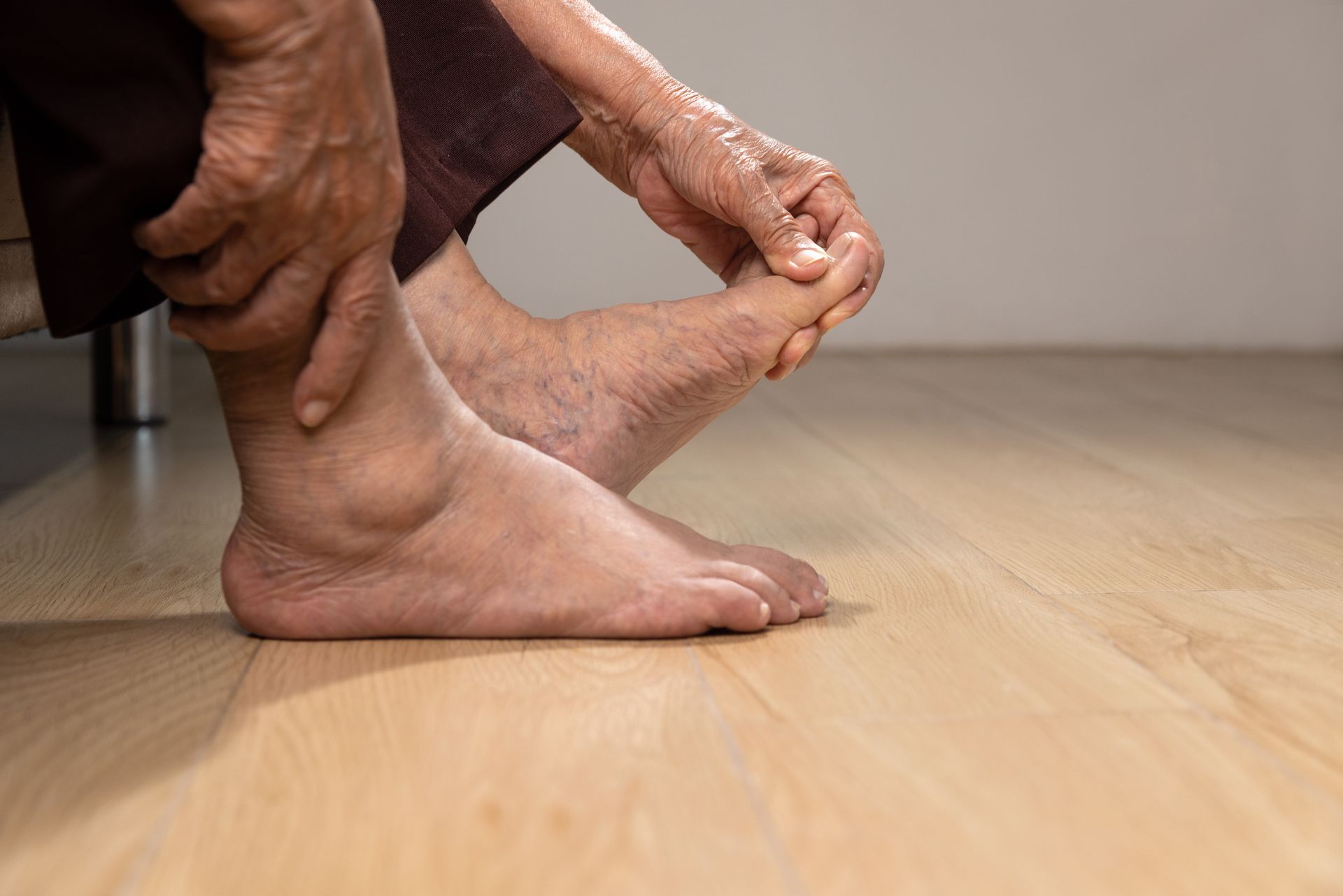 An elderly person holding their swollen foot and ankle, indoors on wood flooring.