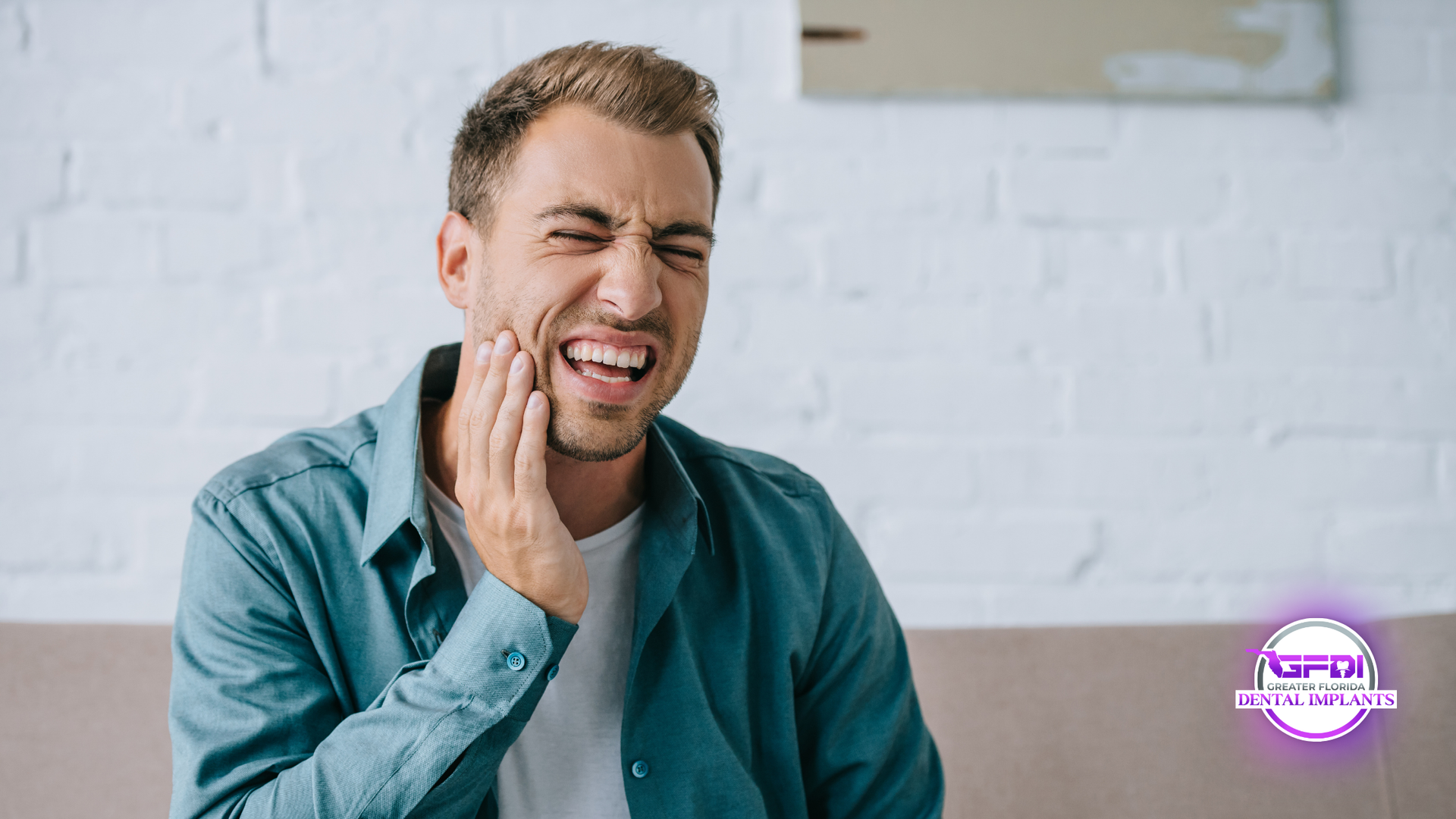 Man with a grimacing expression clutches his cheek, indicating severe tooth pain; indoors on a sofa.