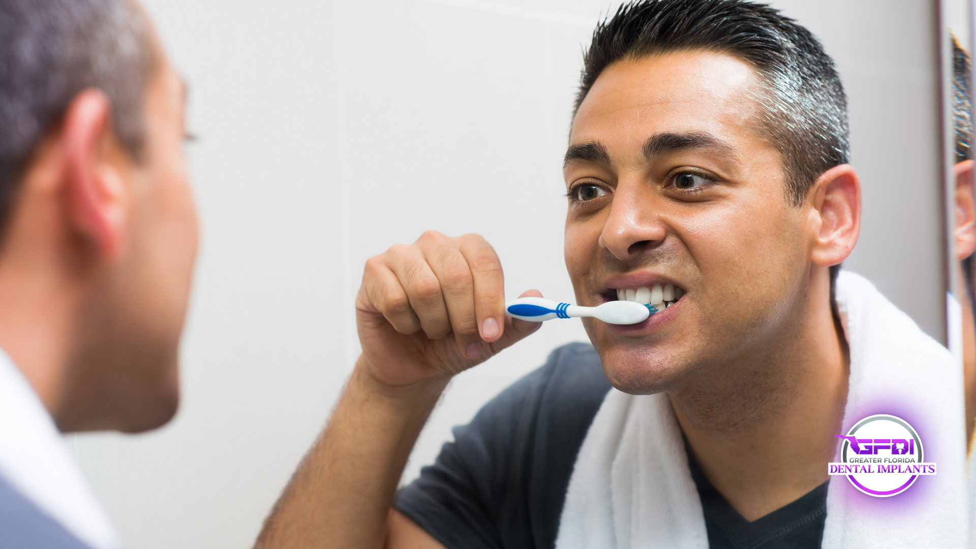 A man brushing his teeth in front of a mirror, smiling. He's holding a toothbrush, wearing a gray shirt and a white towel.