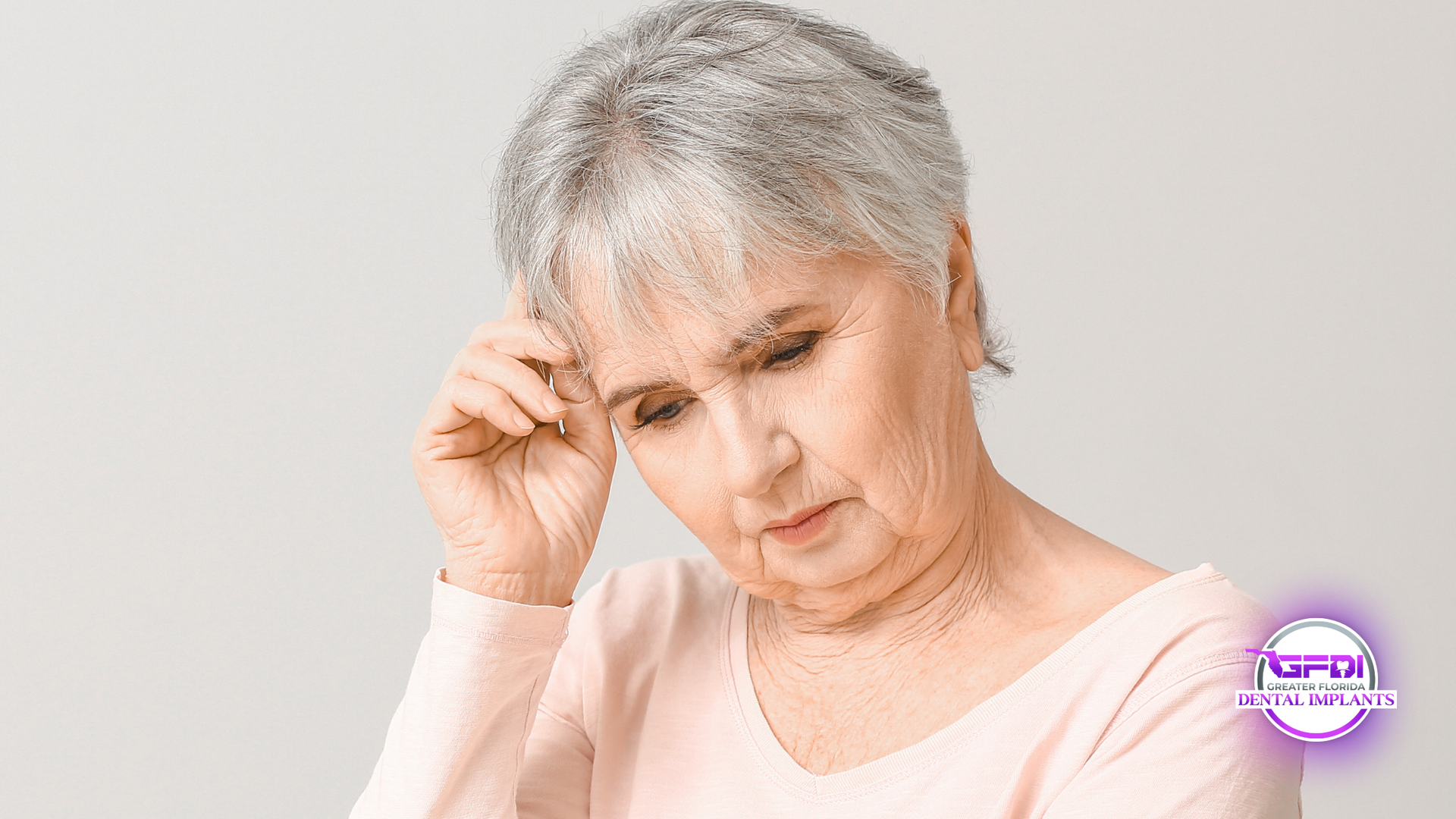 Woman with short gray hair, hand on forehead, looking down, concerned expression, light background.