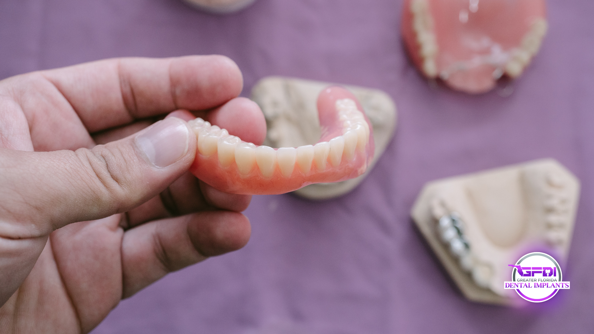 Hand holding upper denture with others on a purple surface.