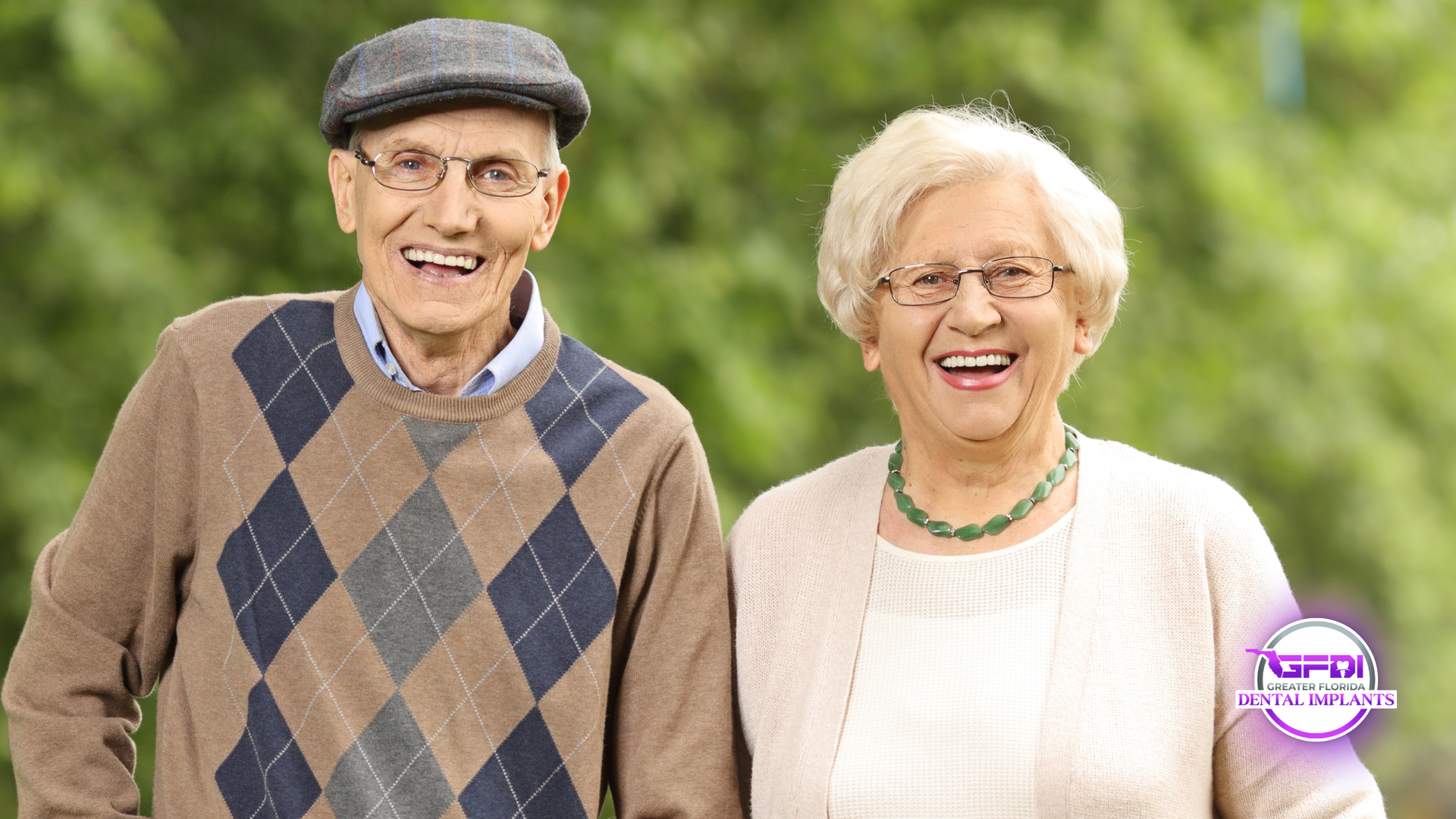 Elderly couple smiling outdoors. Man wears a hat and sweater, woman wears glasses and a necklace, both happy.