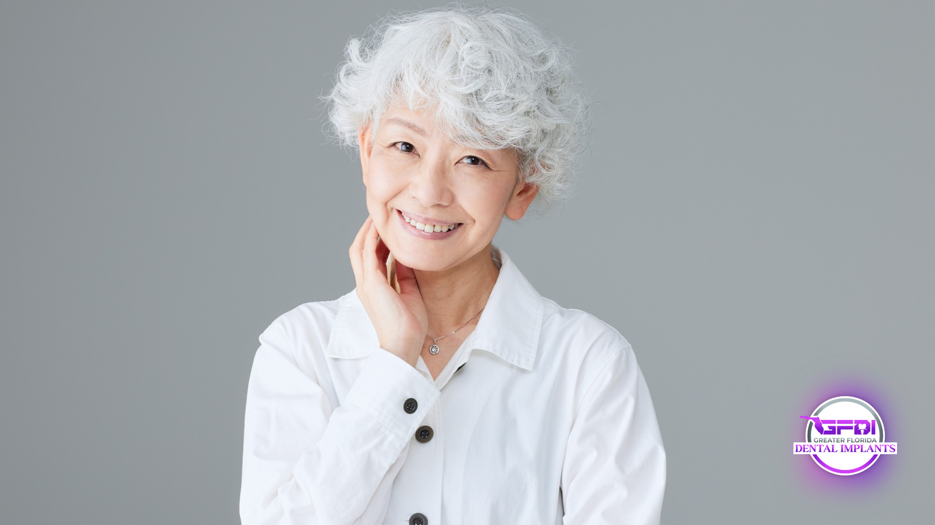 Woman with curly, gray hair smiling, wearing a white shirt, hand near her neck.