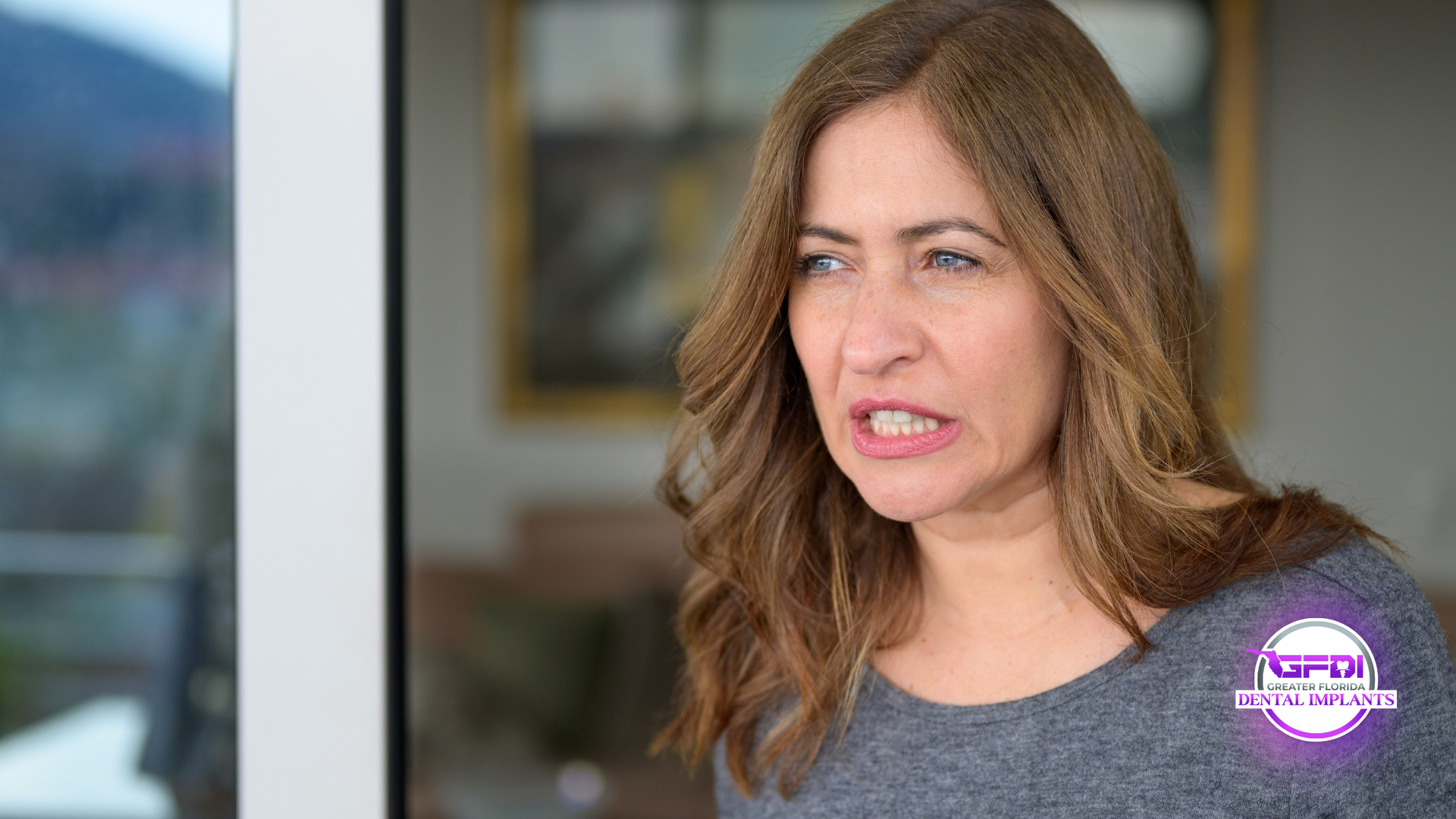 Woman with brown hair makes a disgusted face, standing near a glass door with a blurred outdoor view.
