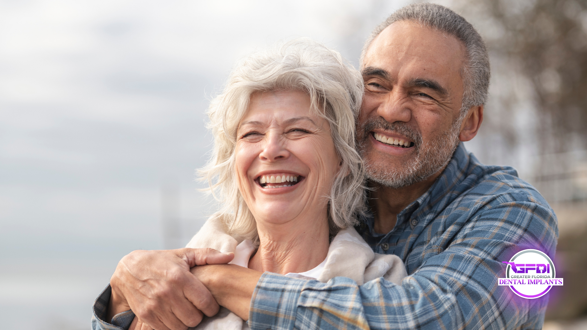 Smiling elderly couple embracing outdoors. Woman laughs with man's arms around her, against a blurred sky.