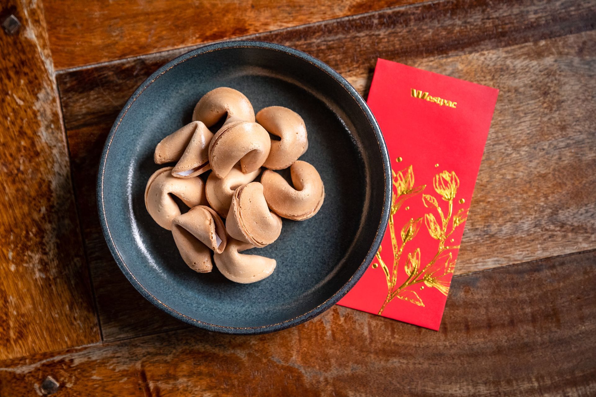 A bowl of fortune cookies next to a red envelope on a wooden table.