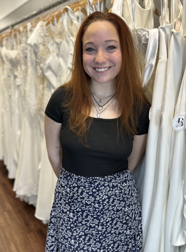 Woman smiling, standing in a bridal shop, wearing a black shirt and floral skirt, surrounded by wedding dresses.