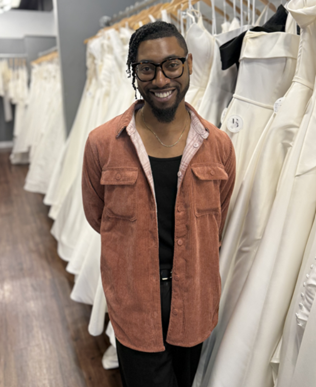 Man smiling, standing between rows of wedding dresses. He wears a corduroy shirt, black top, and glasses.