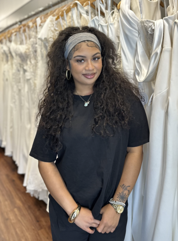 Woman in black t-shirt and headband smiles in a bridal shop, dresses in the background.
