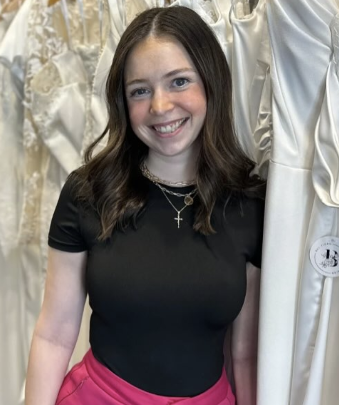 Woman smiles in a wedding dress shop, wearing a black top and pink pants.