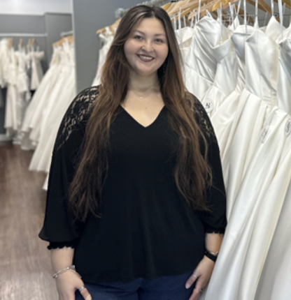 Woman smiling in a bridal shop, wearing a black top with lace sleeves, surrounded by wedding dresses.
