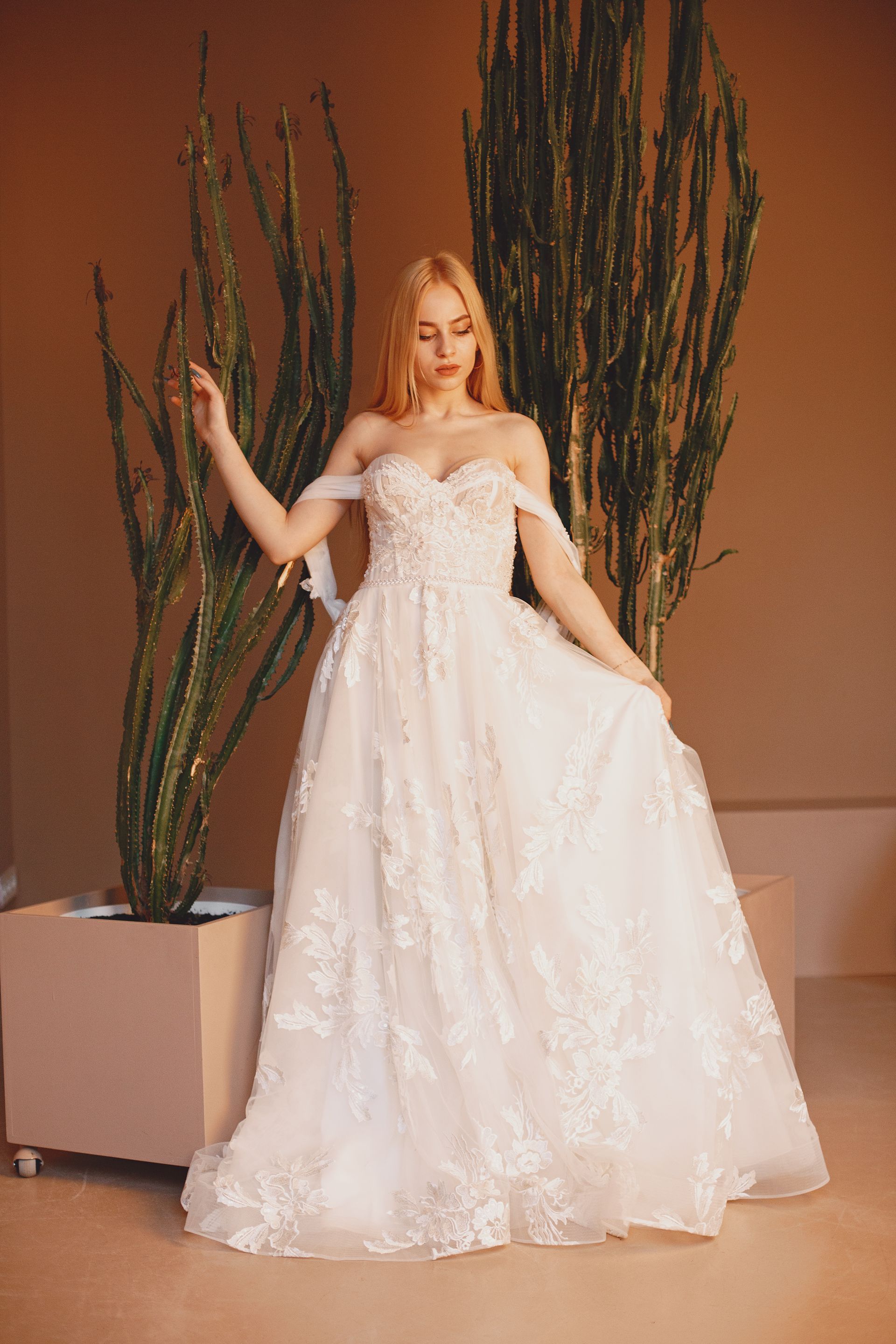 Blonde woman in a white off-shoulder wedding dress poses by a tall cactus in a tan room.