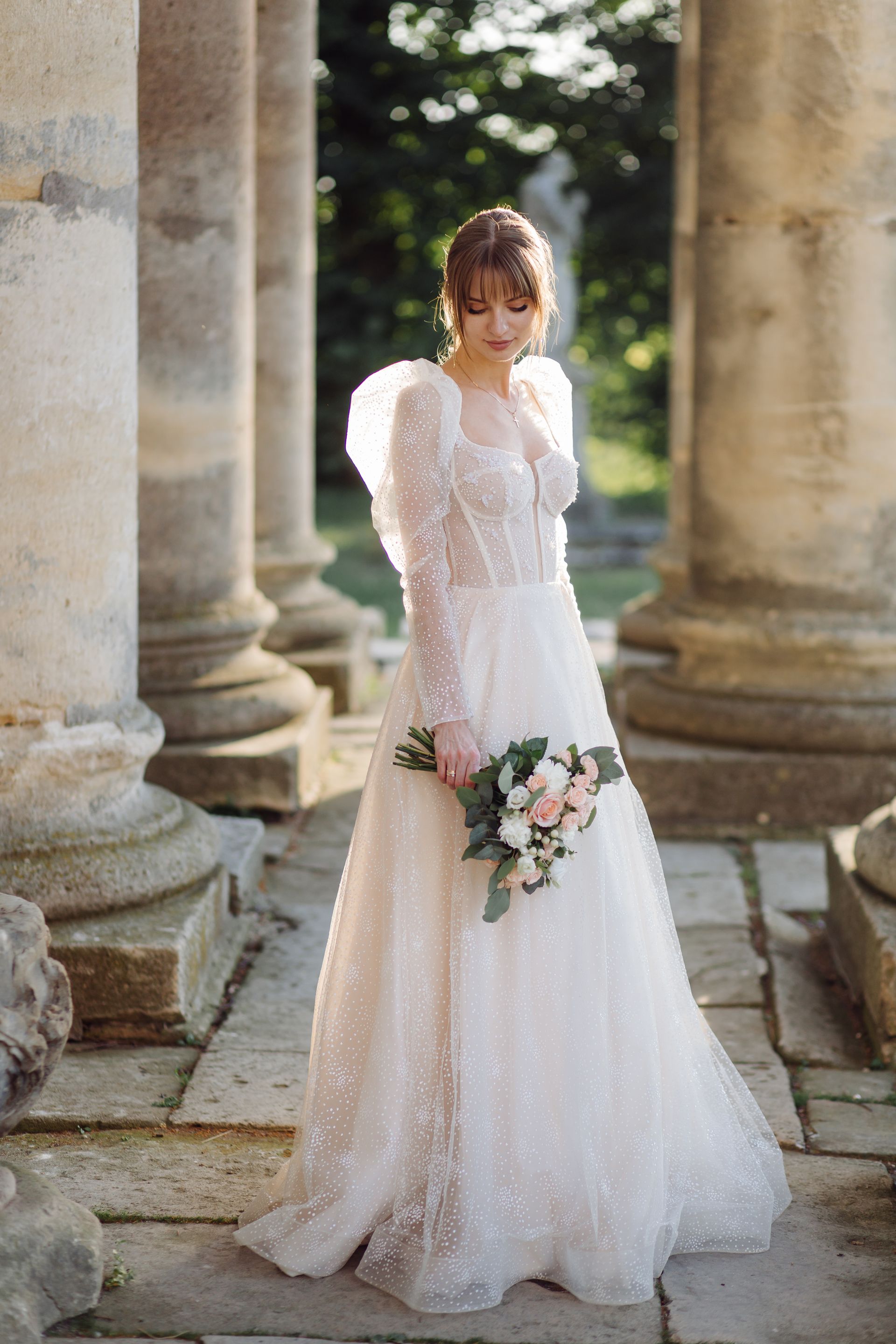 Bride in a sparkling white gown holds flowers, standing near stone columns outside.