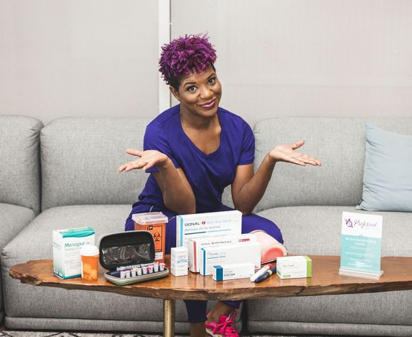 Woman with purple hair seated with medical supplies on a table; she gestures with open hands, smiling.