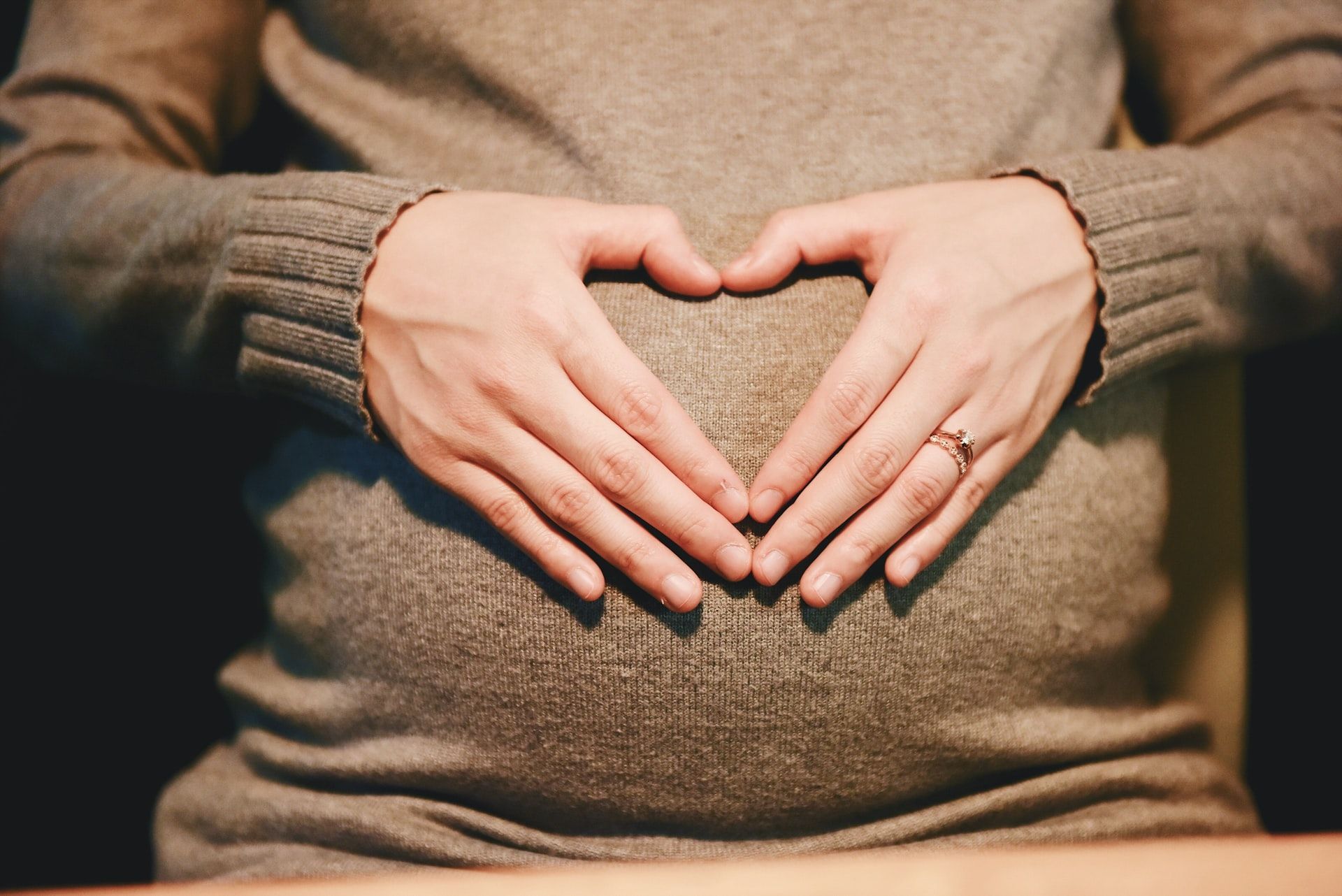 Pregnant person with hands forming a heart shape on their abdomen, wearing a gray sweater.