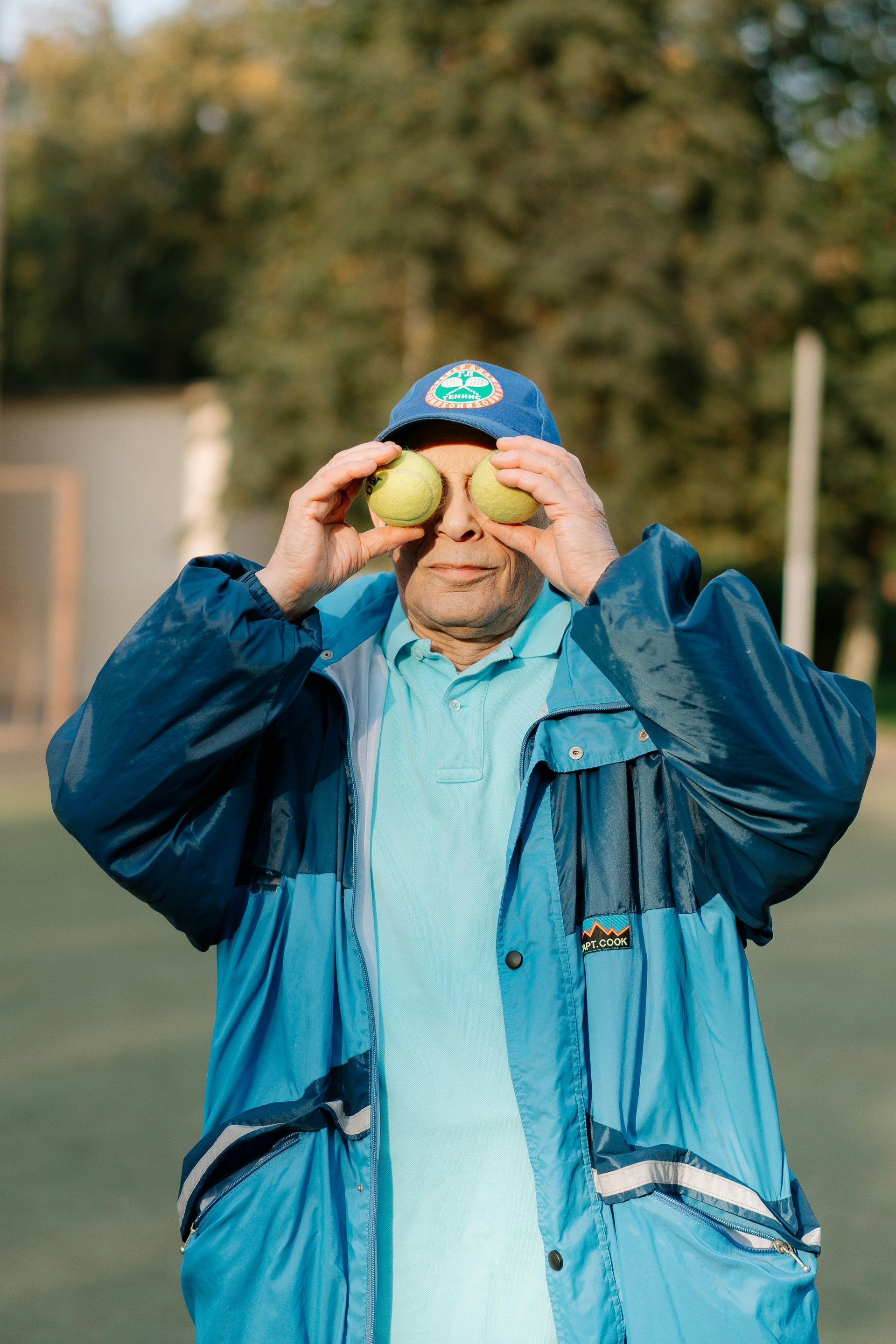 Man holding tennis balls over eyes, wearing blue cap and jacket, on a tennis court.