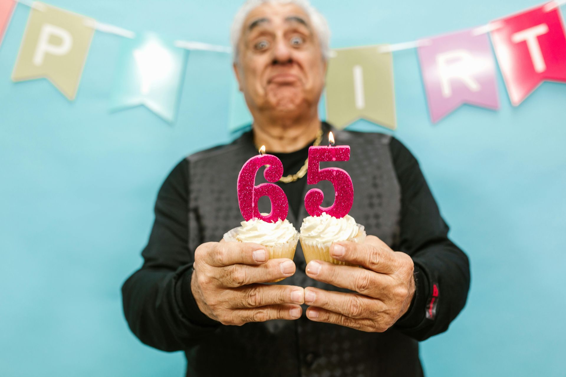 Elderly man with cupcakes, celebrating 65th birthday, against a blue backdrop with birthday banner.