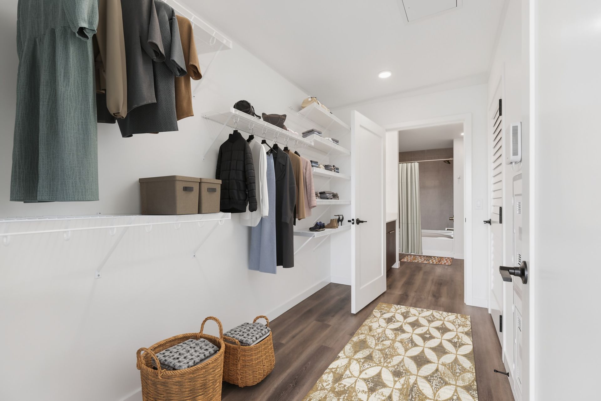 Bright white walk-in closet with hanging clothes, shelves, baskets, and a patterned rug in a hallway