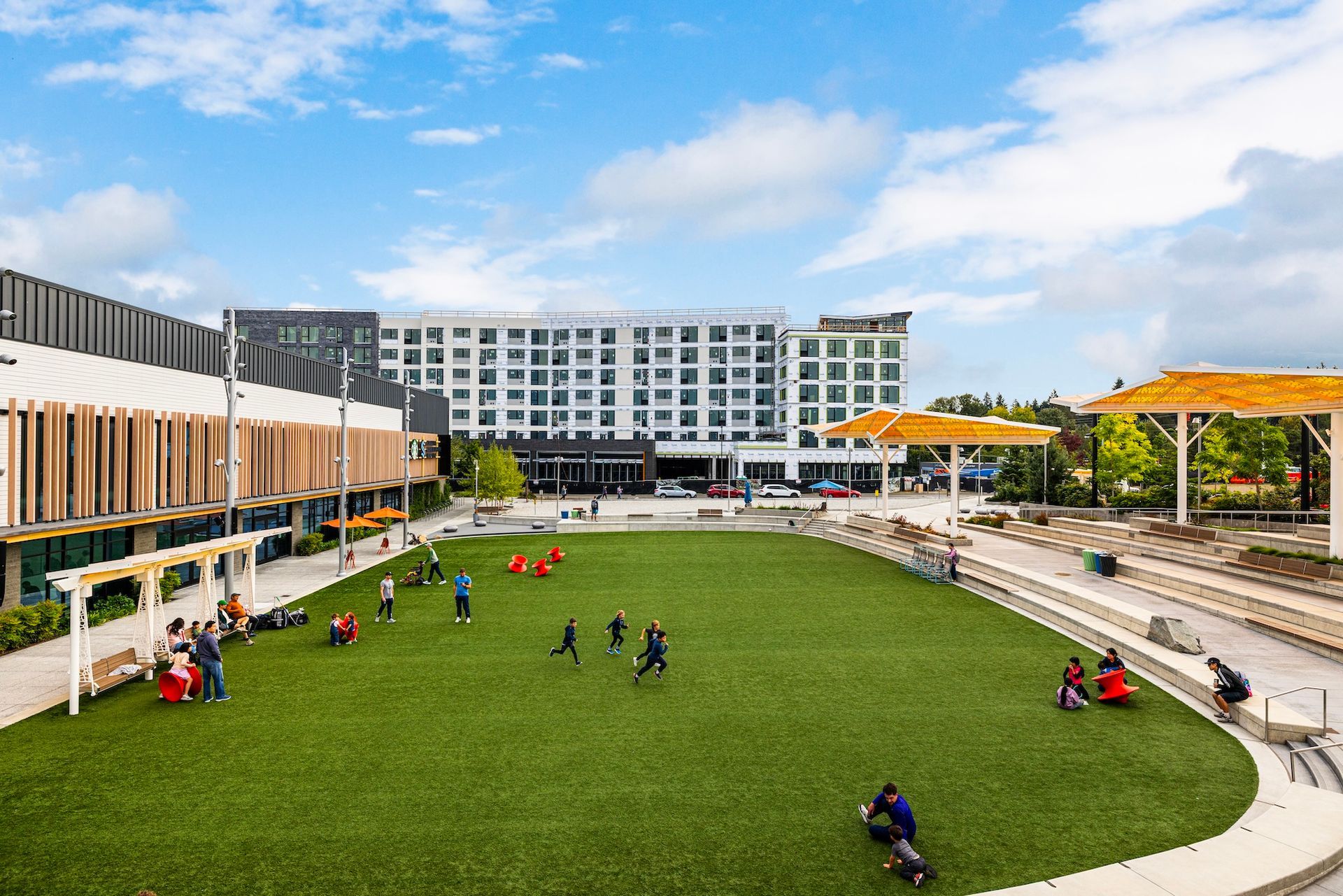 Modern campus courtyard with green lawn, students sitting and walking, flanked by buildings and yellow shade canopies