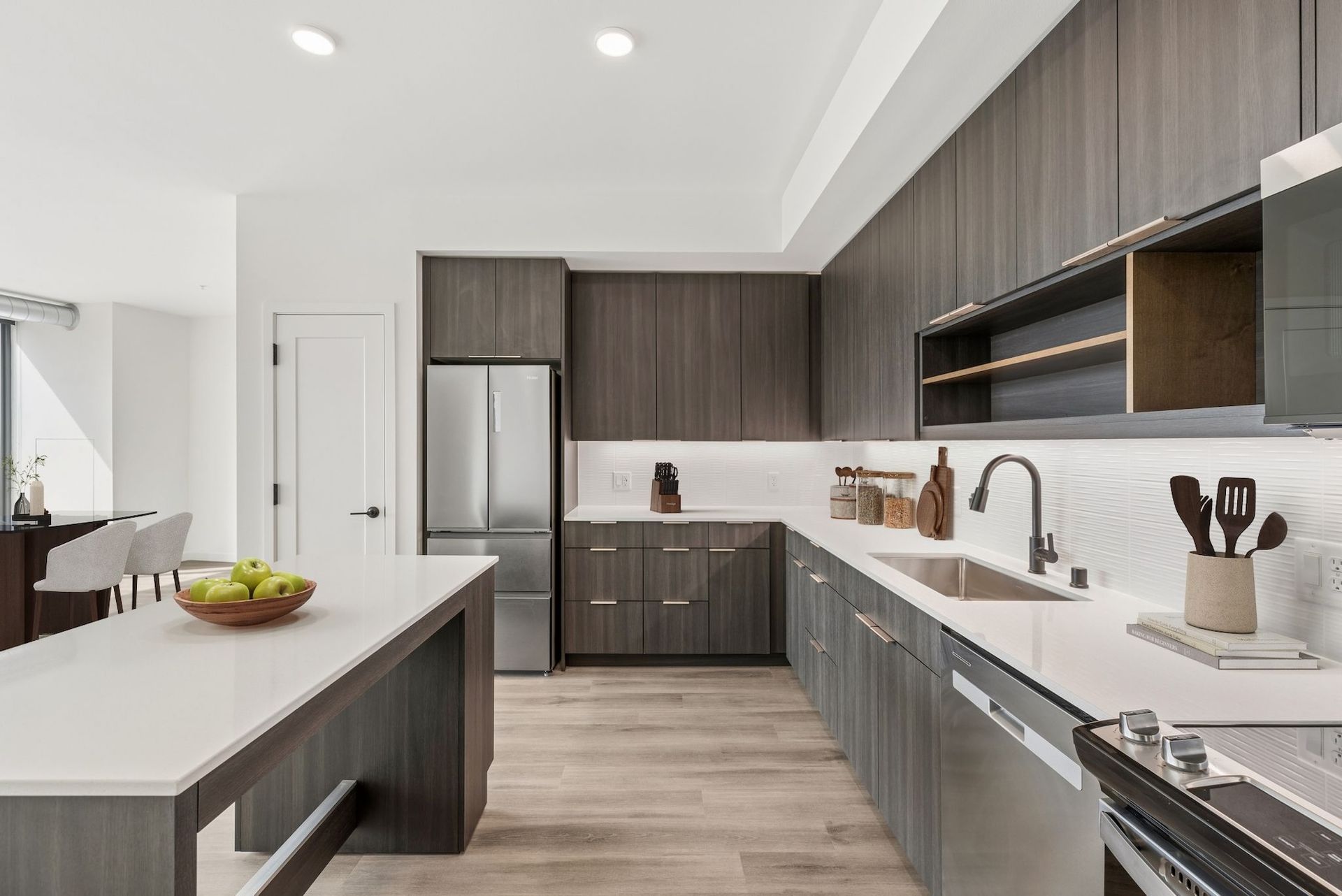 Modern white kitchen with gray wood cabinets, island, stainless steel fridge, and bright recessed lighting
