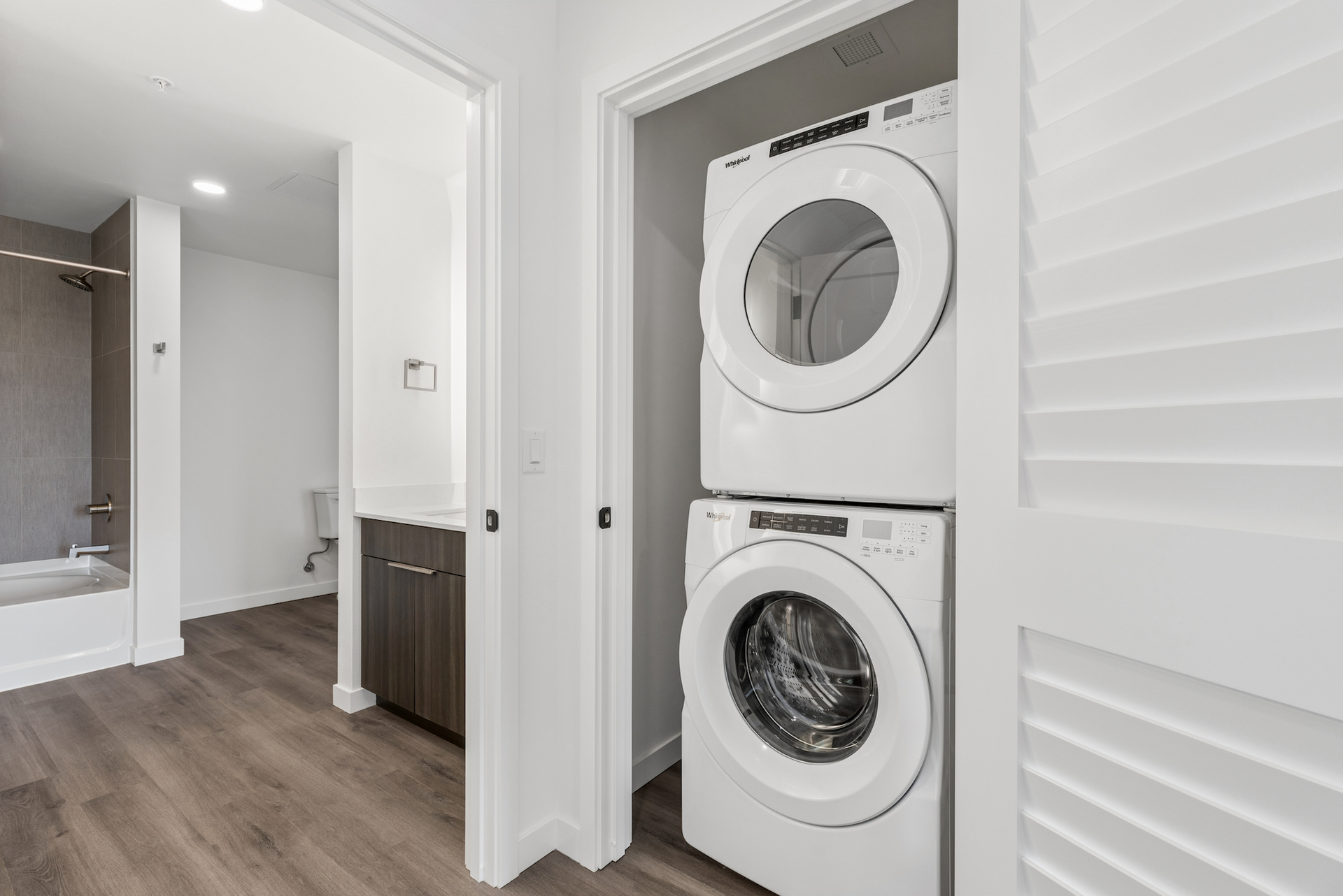 Stacked white washer and dryer in a hallway laundry closet beside a bathroom and wood floor