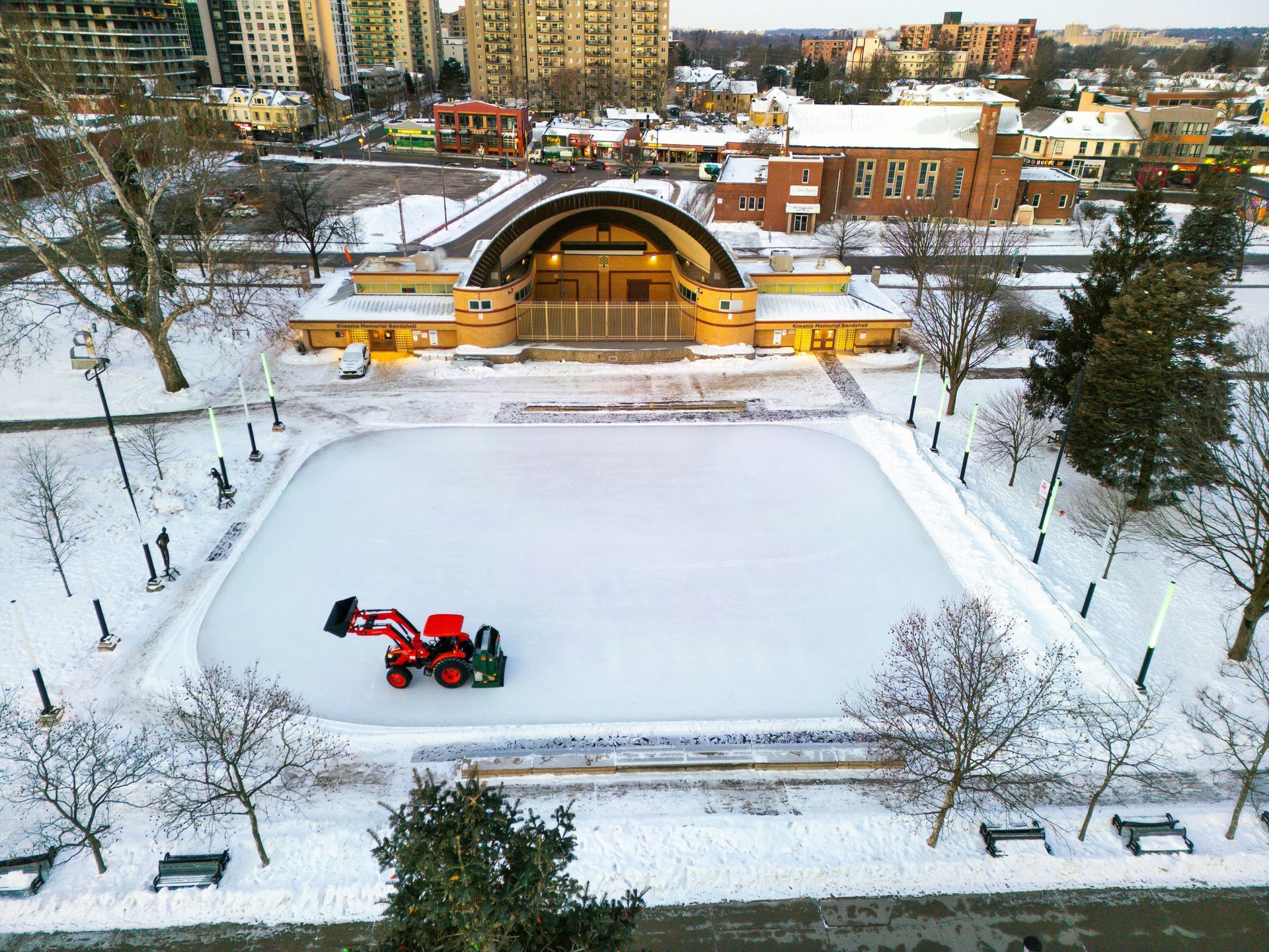Snow-covered city ice rink with a red Zamboni resurfacing the ice near a lit pavilion.
