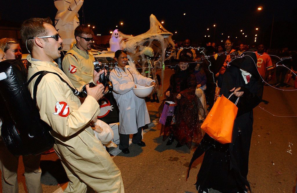 People in Halloween costumes trick-or-treating at night. Ghostbusters, Grim Reaper, and other costumes are visible.