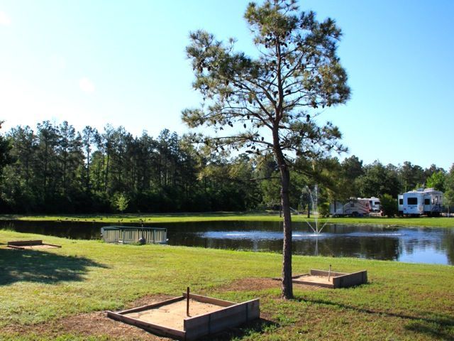 Horseshoe pits on grassy area near pond and trees, sunny day with RVs in the background.
