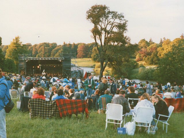 Crowd at an outdoor concert, gathered on a grassy field, stage in background.